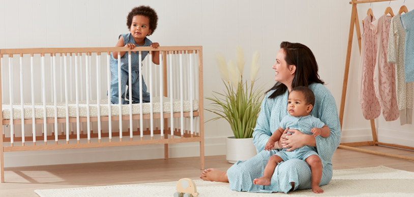 Mom sitting on ground holding baby, looking at toddler in crib