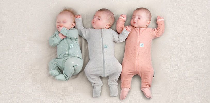 Three newborns sleeping side by side, wearing ergoPouch Rompers in Sage, Grey Marle and Berries