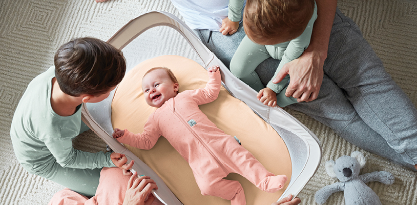 Two young children lean over the Easy Sleep Portable Baby Crib inside of which baby is smiling