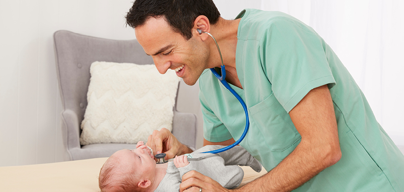 Dr Golly (Dr Daniel Golshevsky) leaning over a baby, listening to heartbeat with stethoscope and smiling