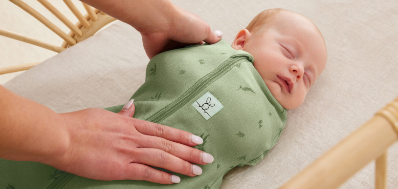 sleeping baby swaddled in green ergoPouch swaddle bag, being settled by a hand in resting in crib